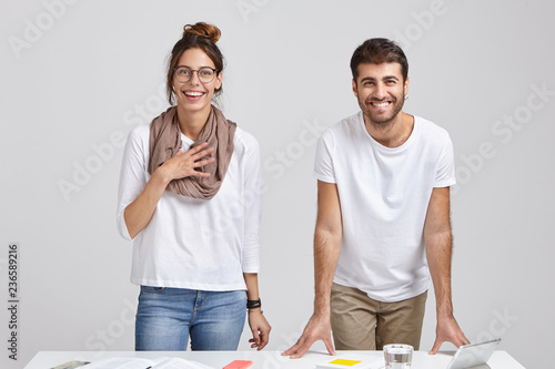Fotografía Shot of happy colleagues rejoice praise from boss for good job, have toothy smiles, stand near desk with tablet, glass of water and books, isolated over white studio background