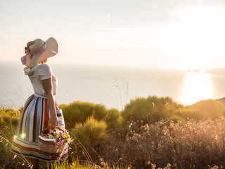 Young woman with flowers basket enjoying sunset view on the hill over the seaside. Large hat, fashion white shirt, colorful skirt.