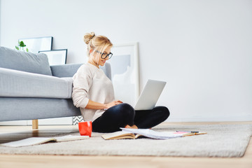 Female student sitting on floor of her apartment with laptop and notes studying