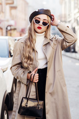 Wall Mural - Outdoor fashion portrait of young beautiful woman wearing trendy beige trench coat, stylish cat eye sunglasses, leather beret, turtleneck, holding small bag, model posing in street of european city