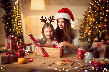 Happy mother in santa helper hat and little girl in Christmas deer antlers pack Christmas gifts and have fun over living room, christmas tree, lights background