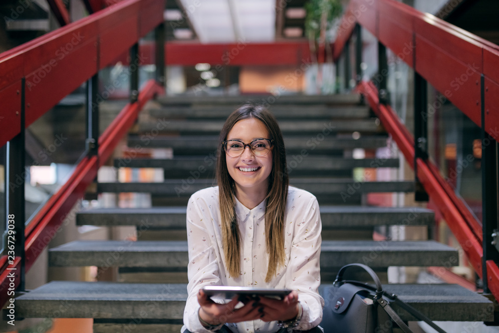 Young Caucasian female student with brown hair and eyeglasses using tablet while sitting on the stairs in college building. Next to her bag. - obrazy, fototapety, plakaty 