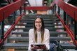 © dusanpetkovic1 - Young Caucasian female student with brown hair and eyeglasses using tablet while sitting on the stairs in college building. Next to her bag.