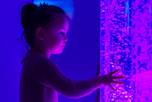 Child In Therapy Sensory Stimulating Room, Snoezelen. Child Interacting With Colored Lights Bubble Tube Lamp During Therapy Session.