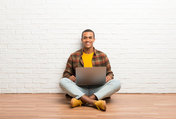 Wall Mural - African american man sitting on the floor with his laptop posing with arms at hip and laughing looking to the front