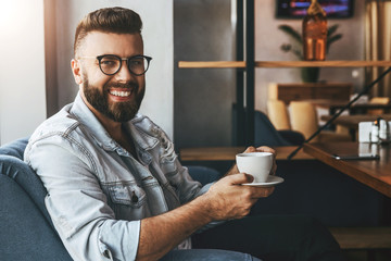 Young attractive bearded businessman in trendy glasses sits in cafe, drinks coffee during lunch time, rests, enjoys drink. Hipster guy waiting for friends in coffee shop. Lifestyle, leisure, pastime.