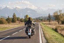 Back View Of Biker In Black Leather Jacket Riding Motorcycle Along Road On Blurred Background Of Beautiful Mountain Range With Snowy Peaks, Moving Vehicles On Bright Sunny Summer Day.