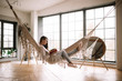 Leinwandbild Motiv Dark-haired girl dressed in pants, sweater and warm slippers reads a book lying in a hammock in a cozy room with wooden floor and panoramic windows and a round mirror on the floor