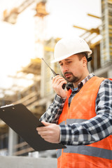 Developing engineer wearing white safety vest and hardhat with walkie talkie and clipboard inspecting construction site