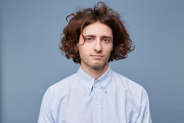 Young guy in a light shirt, with messy untidy hairstyle, long brown hair blue eyes, looking calmly into the camera standing against blue background.