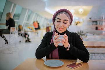 Wall Mural - Attractive young arabian woman sit at table in cafe. She holds cup in hand and look at it. She smiles.