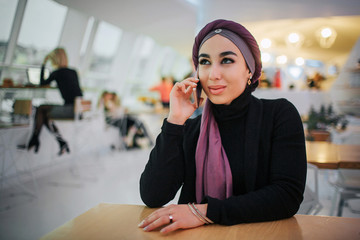 Wall Mural - Attractive young arabian woman sit at table in cafe. She talks on phone. Model smiles. She wears hijab.