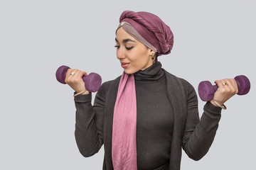 Wall Mural - Strong young arabian woman exercising with dumbbells. She looks at one of it. She wears hijab. Isolated on grey background.