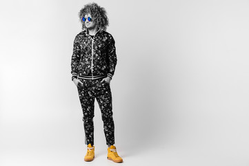 Wall Mural - Adult shylish man in fashionable tracksuit with flowers pattern, yellow sneakers standing with hands in pockets on white background. Guy with curly hair. Awesome confident male in sunglasses portrait