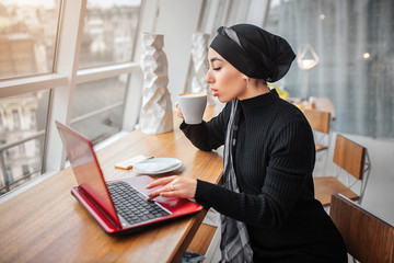 Wall Mural - Beautiful and well-dressed young arabian woman drinks coffee and works on laptop. She sits at table inside. It is sunny outside.