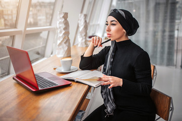 Wall Mural - Thoughtful young man sit at table in cafe near window. She looks at it. Arabian woman hold pen in hand. There are notebook. laptop and cup of coffee on table.