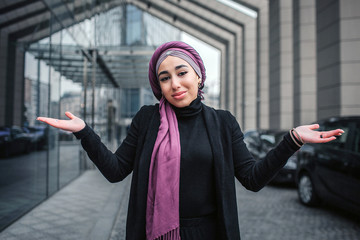 Wall Mural - Happy arabian woman stand outside and look on camera. She holds hands aside of body. Woman wear black clothes and violet hijab.