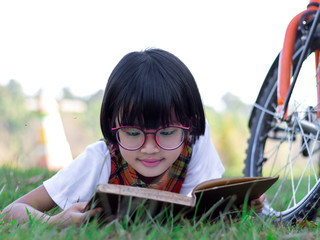 Wall Mural - A little cute girl with glasses  reading a book lying on the grasses in garden at sunset time