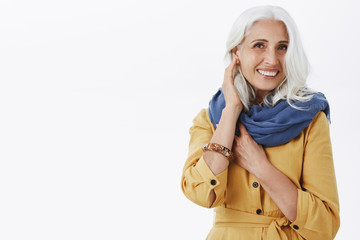 How do i look. Charming fashionable and pleased grandmother with pretty grey hairstyle in trendy yellow coat and blue scarf touching hair gently and smiling at camera asking advice about outfit