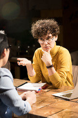 Serious confused confident business lady with curly hair sitting at table and gesturing hand while asking questions to colleague at informal meeting in dark office