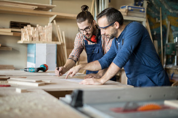 Portrait of two carpenter discussing project standing at table and drawing plans in workshop, copy space