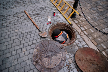 Working man comes out from the sewerage hatch in the ground on city street. Repair of sewage or underground utilities, nightman cleans drains, cable laying. Worker on street cleaning pipe.