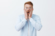 © Cookie Studio - Man being shocked witnessing terrible scene. Portrait of stunned terrified redhead male in glasses and shirts dropping jaw, gasping from amazement, staring at camera worried over gray background