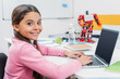 © LIGHTFIELD STUDIOS - adorable schoolgirl sitting at table with robot model, looking at camera and using laptop with blank screen during STEM lesson