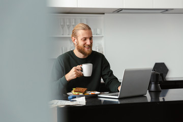 Handsome young bearded man sitting at the kitchen drinking coffee using laptop computer.