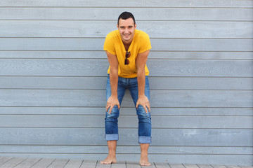 Full body barefoot man smiling against gray wall with hands on knees