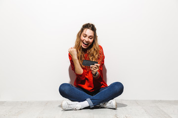 Sticker - Photo of optimistic woman 20s smiling and playing video games on smartphone while sitting on floor with legs crossed, isolated over white background