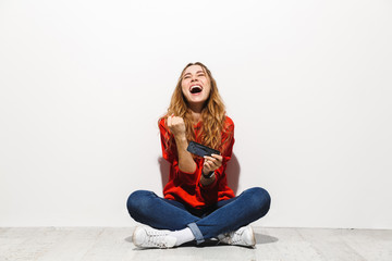 Sticker - Photo of excited woman 20s smiling and playing video games on smartphone while sitting on floor with legs crossed, isolated over white background