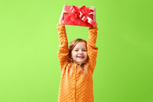Portrait Of A Little Girl In A Warm Knitted Sweater On A Ufo Green Background. A Child Holds A Red Box With A Gift Above His Head. The Concept Of Celebration, Giving And Receiving A Gift.