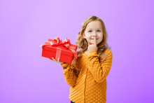 Portrait Of A Little Girl In A Warm Sweater On A Proton Purple Background. The Child Looks Dreamily Up And Holds A Red Box With A Gift. The Concept Of Celebration, Giving And Receiving A Gift