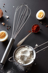 Wall Mural - Sieve with flour, spoon with red pepper, a metal whisk and halves of a boiled egg on a black concrete background. Concept preparation. Flat lay