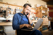 Portrait Of Mature Worker Using Laptop And Speaking By Smartphone  In Workshop Interior, Copy Space