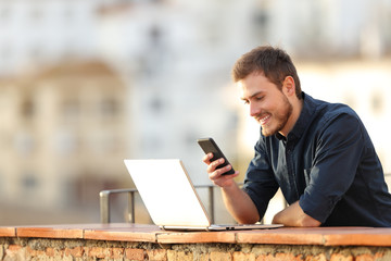 Happy man using phone and laptop in a balcony