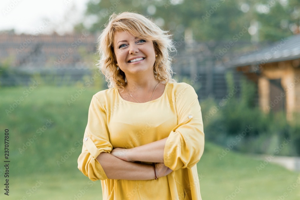 Outdoor portrait of positive confident mature woman. Smiling female blonde in a yellow dress with arms crossed near the house - obrazy, fototapety, plakaty 