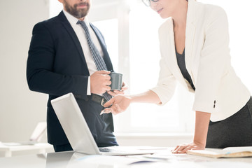 Busy colleagues standing at table and discussing online data on laptop: businesswoman in jacket pointing at screen while explaining information to man with mug