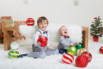 Portrait of white Caucasian children brothers sitting together near wooden bench smiling, celebrating Christmas or New Year. Little cute boys siblings playing in studio with holiday decoration.