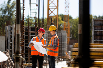 Construction manager and engineer dressed in orange work vests and hard helmets explore construction documentation on the building site near the steel frames