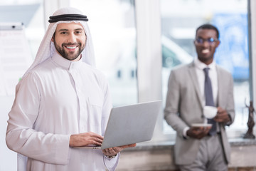 Wall Mural - Arabian businessman holding laptop in office with african american partner on background