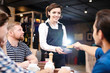 © Seventyfour - Happy positive young lady waitress with short hair standing at table and holding card reader while guest paying for dinner with friends using credit card in restaurant.