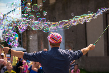 Wall Mural - A freelance clown blowing hundreds of tiny, small and big bubbles at outdoor festival in city center. Concept of entertainment, birthdays. Kids having fun. Shower of bubbles flying in the happy crowd