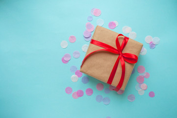 Wall Mural - Gift box wrapped in brown colored craft paper and tied with red bow on blue background with colofrul confetti and copyspace.