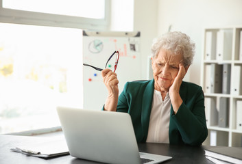 Senior woman suffering from headache while working with laptop indoors