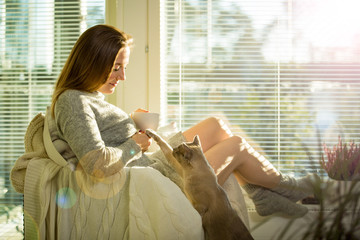 Wall Mural - Woman sitting at home in a chair by the window with cup of hot coffee wearing knitted warm sweater, stroking cat on her laps. Cozy sunny room filled with light. beautiful morning behind big window.