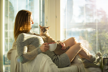 Wall Mural - Woman sitting at home in a chair by the window with cup of hot coffee wearing knitted warm sweater, stroking cat on her laps. Cozy sunny room filled with light. beautiful morning behind big window.