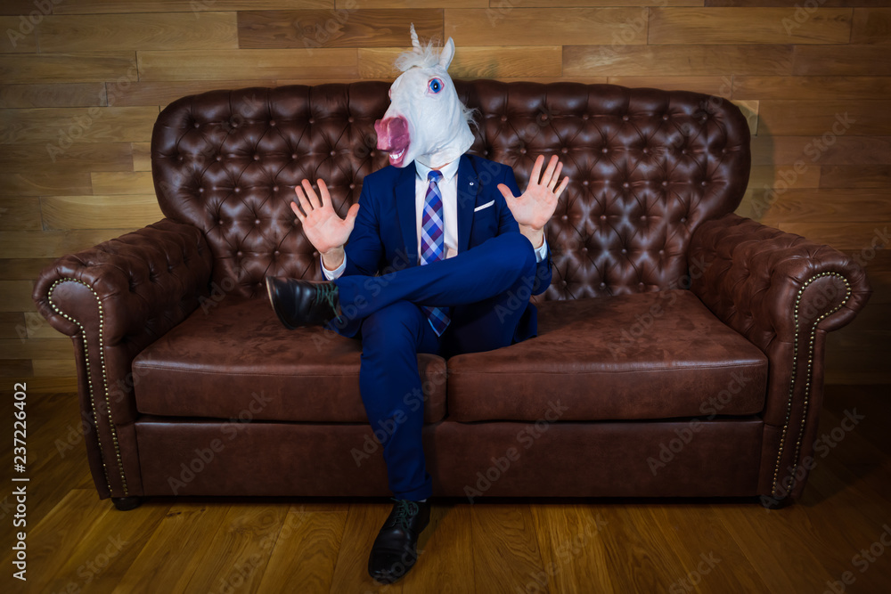 Funny unicorn in elegant suit siting on sofa like a boss and showing do not bother me gesture. Portrait of unusual man at home. Freaky guy in mask. - obrazy, fototapety, plakaty 