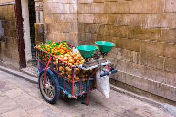 A cart by a Moroccan street fruit vendor equipped with scales for weighing fruits, filled with tangerines and grenades on a narrow medina street in the city of Fez, Morocco